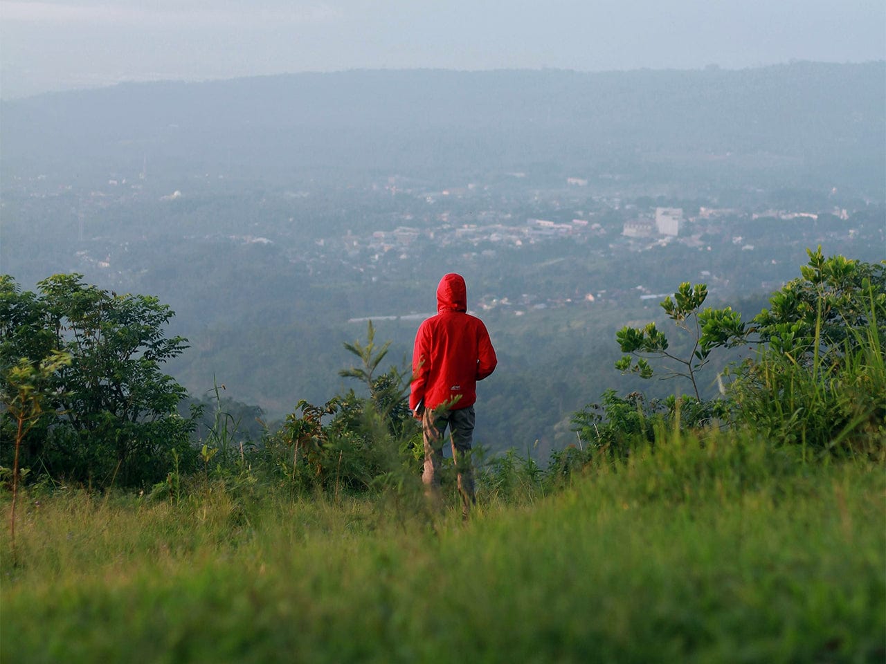Person in a red hoodie standing on a grassy hill overlooking a valley.