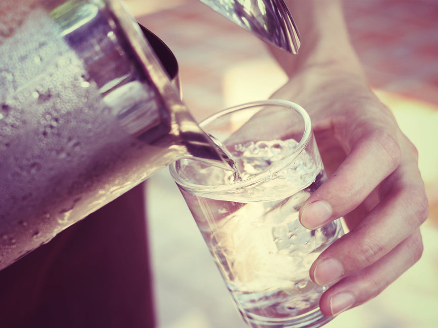 A glass of ice water poured from a silver picture with a woman’s hand holding the glass