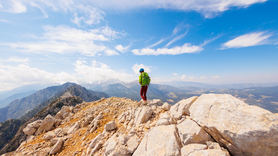 Hiker standing on a rocky mountain summit under a bright blue sky with wispy clouds, looking out over distant mountain ranges and valleys.