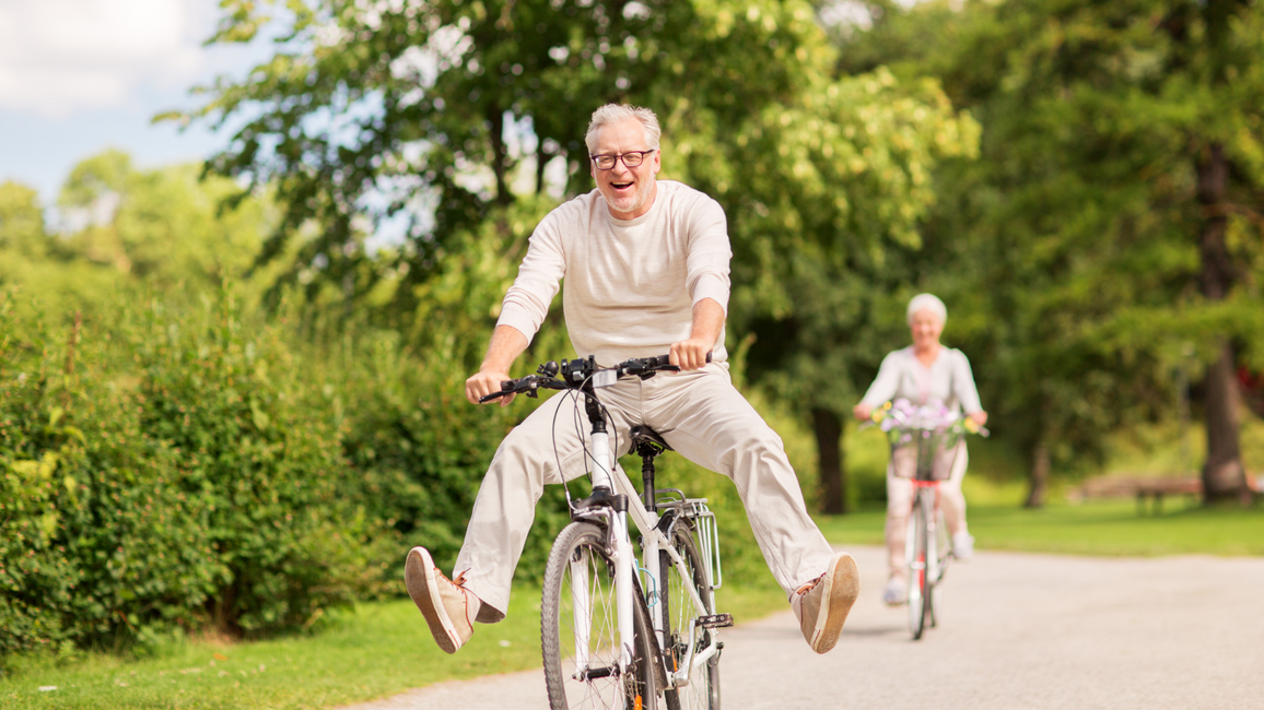 Older man laughing while riding a bicycle down a sunny park path, with another cyclist riding behind him.