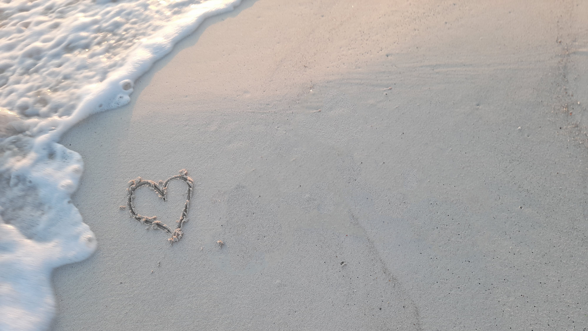 Heart shape drawn in wet sand near the shoreline as gentle ocean foam washes up along the beach.