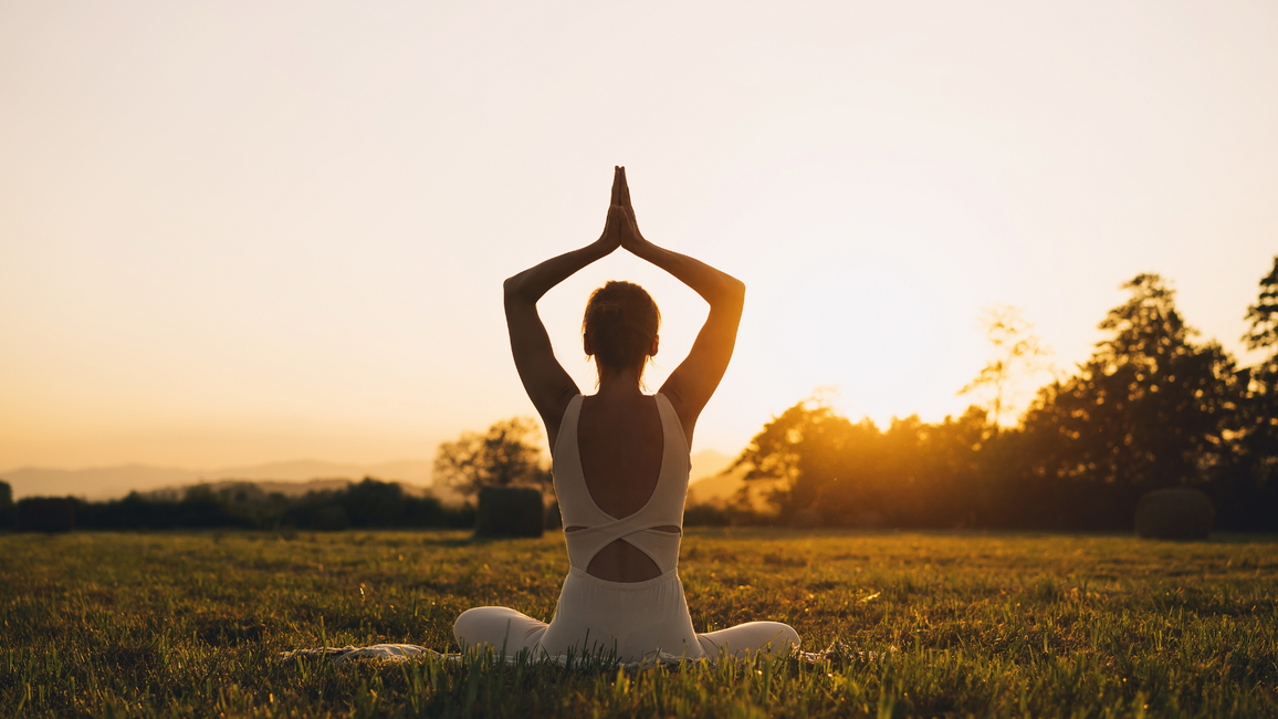Person sitting cross-legged in a grassy field at sunrise, hands pressed together overhead in a yoga pose with the sun glowing behind them.