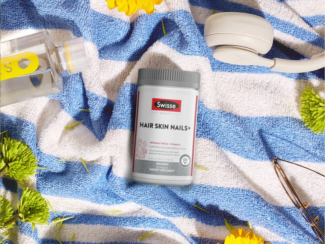 Swisse Beauty Hair Skin Nails+ supplement bottle placed on a blue and white striped towel, surrounded by summer items including headphones, sunglasses, a water bottle, and flowers.