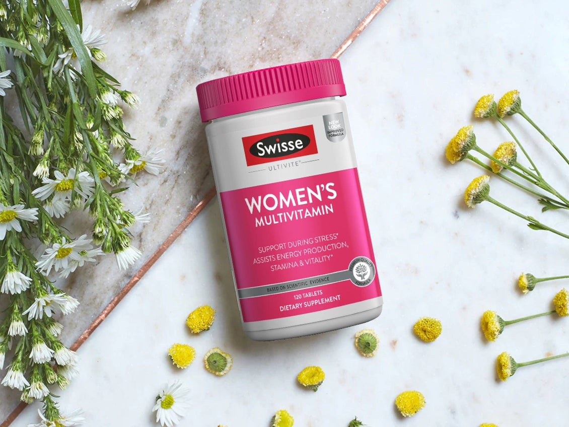 Swisse Women's Multivitamin supplement bottle with a pink label and cap, displayed on a marble surface surrounded by small white and yellow flowers.