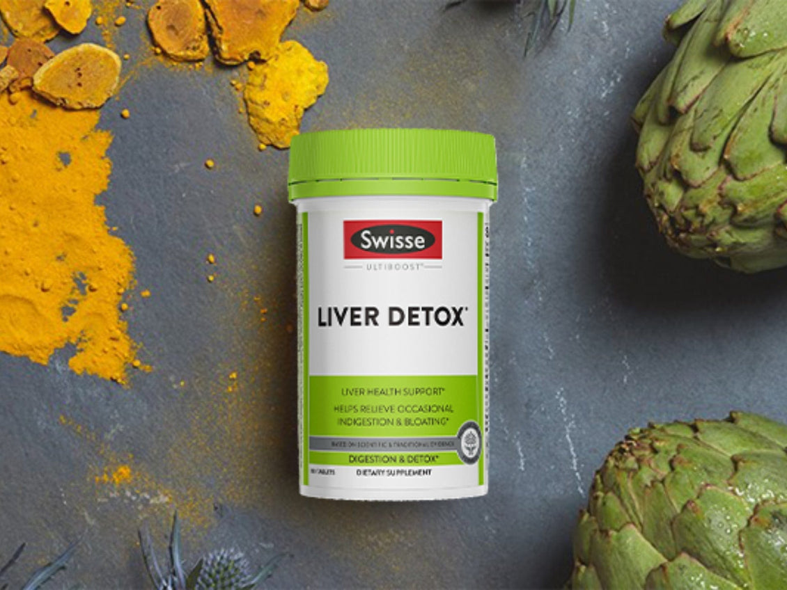 Swisse Ultiboost Liver Detox supplement bottle with green cap, placed on a dark stone surface surrounded by turmeric powder and fresh artichokes.